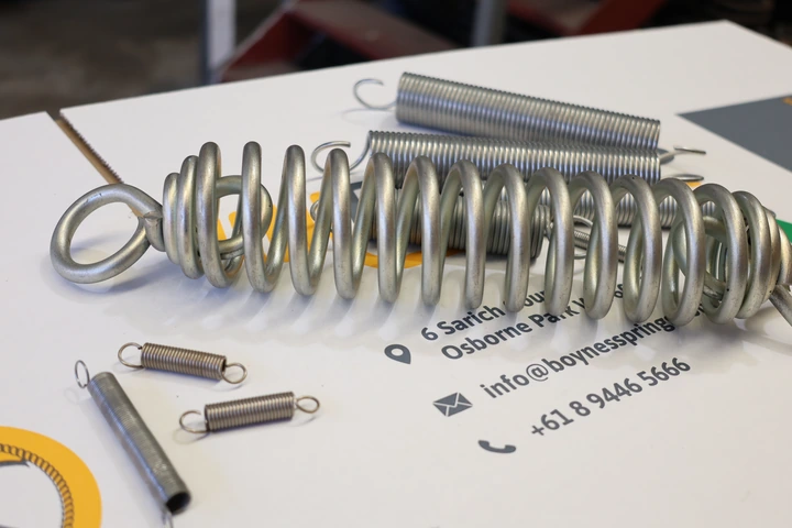 A number of different-sized tension springs, custom made to fit a variety of dimensions and industrial applications, laying on top of the calling card of Boynes Springs, a tension spring supplier in Perth, WA.
